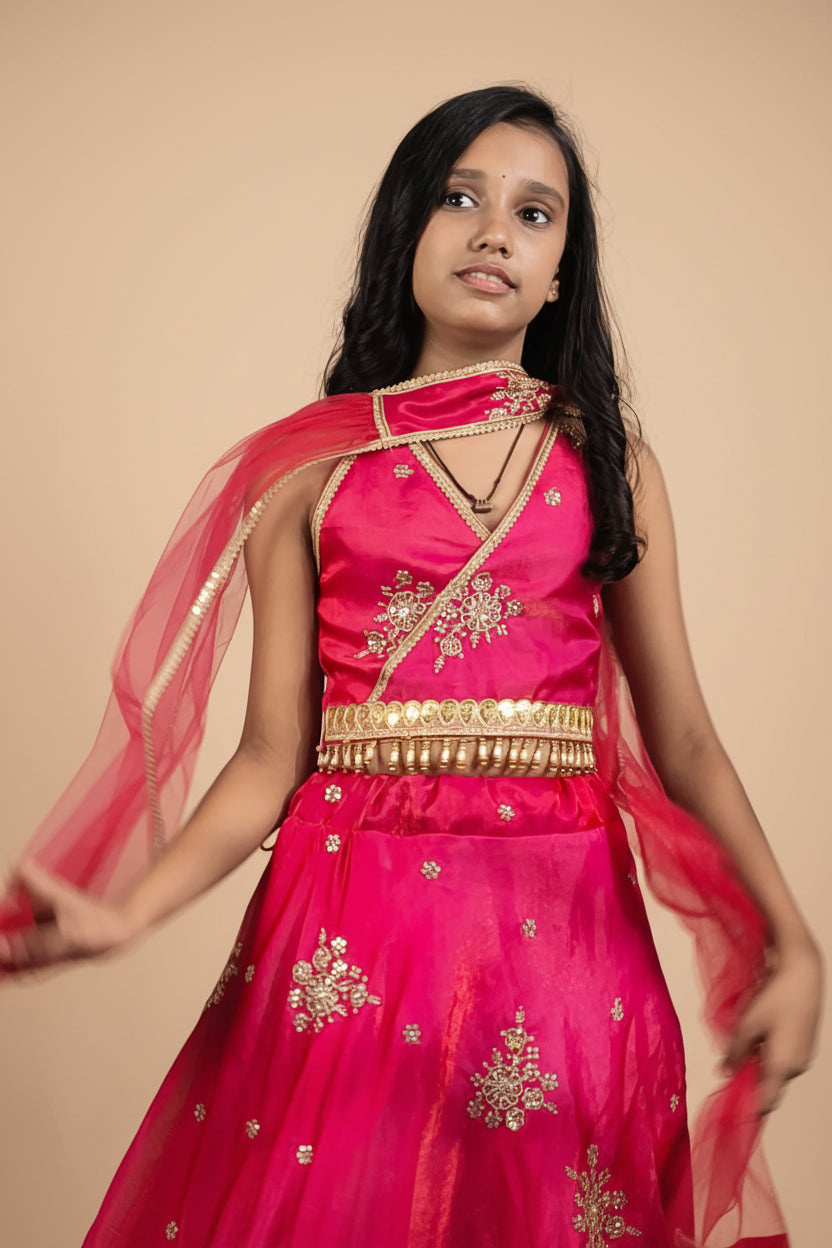 A young girl modeling a bright pink traditional Indian dress with gold embroidery, a gold waist belt, and a sheer pink dupatta, set against a gray studio background with soft leaf shadows.