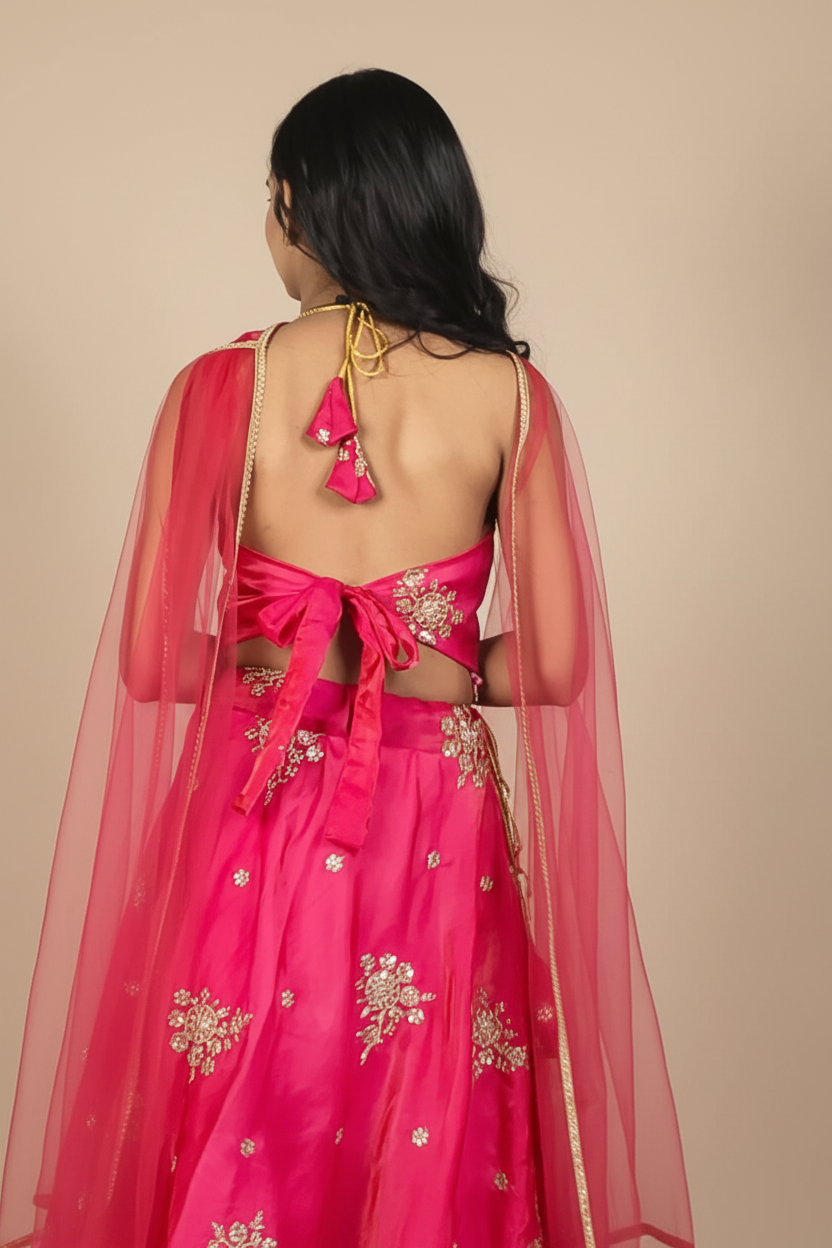 A woman shown from the back wearing a bright pink traditional outfit with gold floral embroidery and a sheer pink dupatta, against a light gray wall with soft shadows.