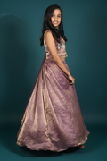 A young girl modeling a traditional Indian ethnic gown with a metallic lavender skirt and a gold embellished crop top, standing against a dark teal studio backdrop.